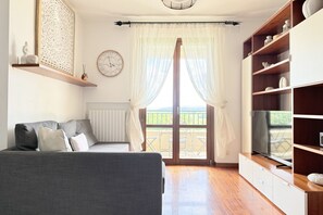 A modern living room with a sofa, a television on a wooden cabinet, a large window with sheer curtains, and a balcony view.