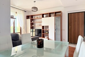 A modern living room with a glass table, white sofa, and a wooden bookshelf.