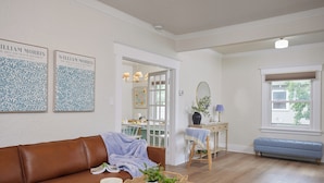 Ein Wohnzimmer mit einem braunen Ledersofa, einem Couchtisch aus Holz mit Kerze und Pflanze, einer blauen Bank und einem Essbereich mit Tisch und Stühlen.