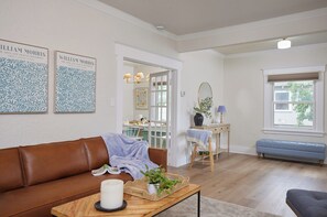A living room with a brown leather couch, a wooden coffee table with a candle and a plant, a blue bench, and a dining area with a table and chairs. - Blue Gables 1920s Craftsman Near WWU 4BR 3BA Sleeps 9 Fenced Yard (College Place)