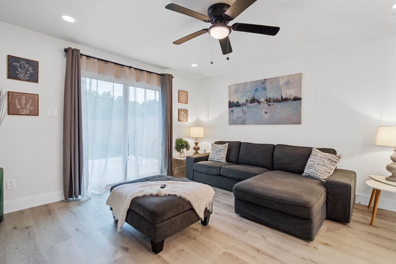 A modern living room with a ceiling fan, a sofa, an ottoman, and a large window with curtains.