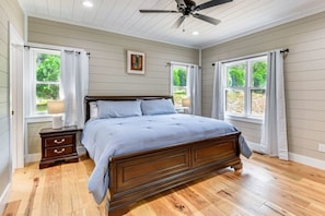 Ein Schlafzimmer mit einem hölzernen Bett, zwei Nachttischen, einem Deckenventilator und großen Fenstern mit Vorhängen.