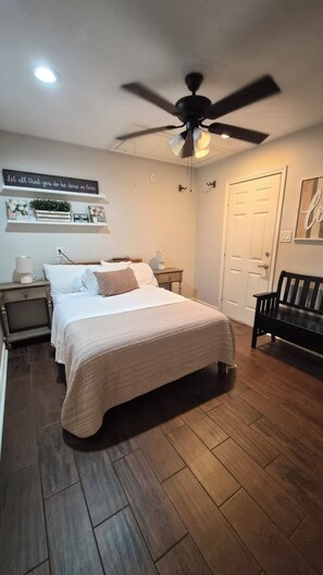 A bedroom with a bed, bedside tables, a ceiling fan, a wall-mounted light, a door, and a bench.