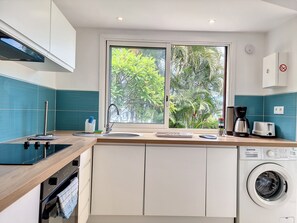 A modern kitchen with a built-in washing machine, a window with a view of greenery, and a coffee maker. - Aybowan - Jacuzzi - Terrasse - AC (Les Avirons)