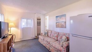 A room with a sofa, a television, a lamp, a refrigerator, and a painting on the wall.
