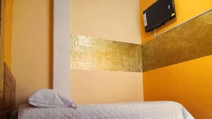 Un lit simple avec une couette blanche, dans une pièce dont le sol est carrelé et où est fixée un téléviseur au mur.