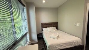 Um quarto com cama, mesa de cabeceira, ventilador de teto e janela com persianas.