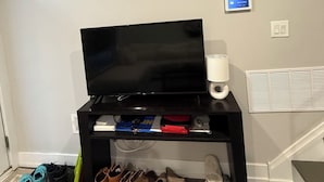 Sala de estar com televisão de tela plana em suporte preto, abajur branco, termostato fixado na parede e sapateira com vários pares de sapatos.