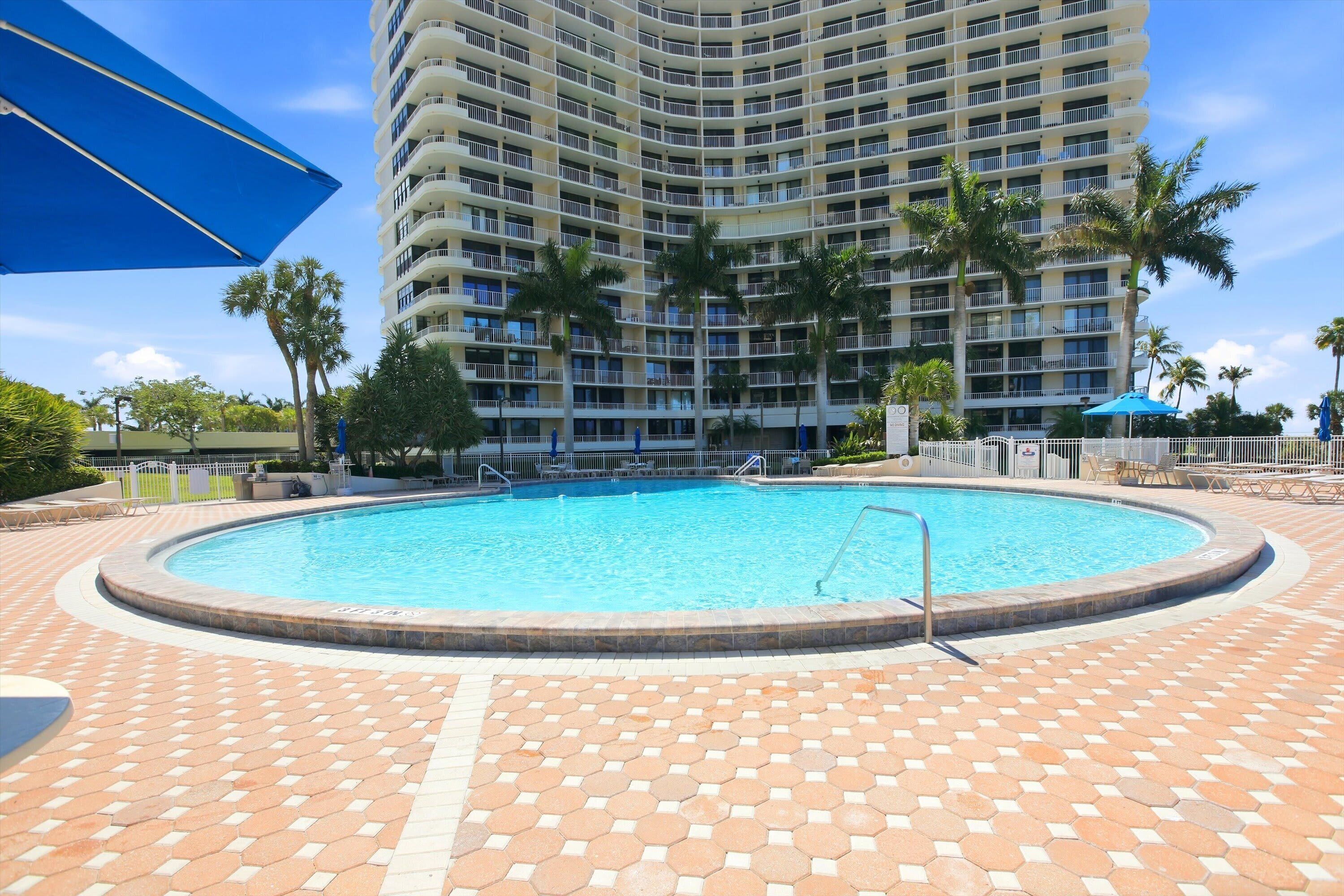 A swimming pool with a tiled deck, surrounded by palm trees and a tall hotel building.
