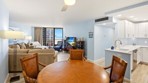Ein modernes Wohnzimmer mit einer Couch, einem Esstisch und Stühlen. Es gibt eine Küchenzeile mit weißen Schränken und einer Spüle. Ein Deckenventilator ist sichtbar, und ein Fenster mit Vorhängen ist vorhanden.