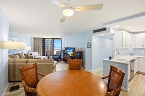 Ein modernes Wohnzimmer mit einer Couch, einem Esstisch und Stühlen. Es gibt eine Küchenzeile mit weißen Schränken und einer Spüle. Ein Deckenventilator ist sichtbar, und ein Fenster mit Vorhängen ist vorhanden.