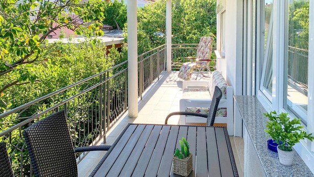 En balkon med fletmøbler, en potteplante og udsigt til grønne områder og bygninger.
