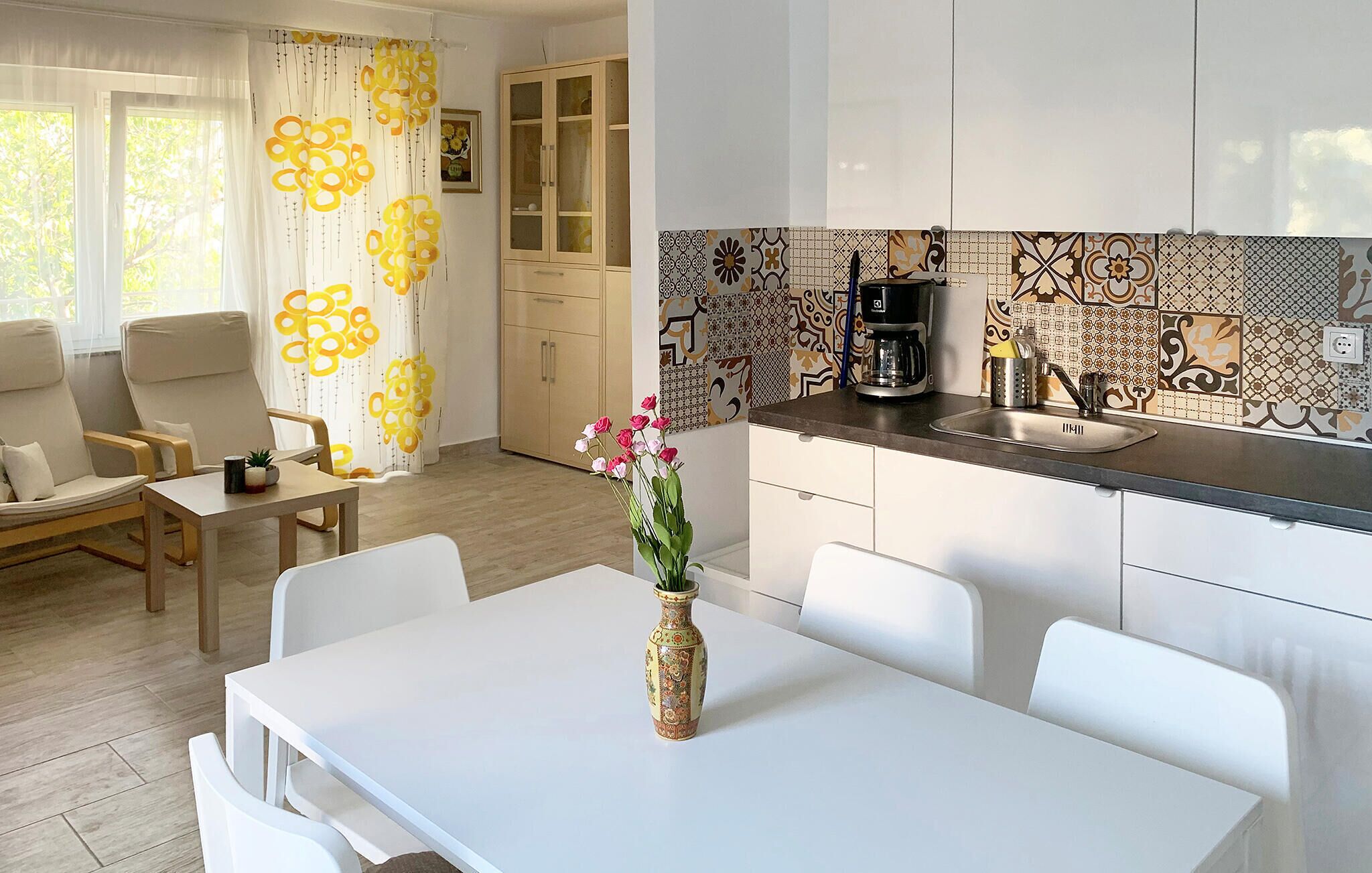 Een moderne keuken met een spoelbak, koffiezetapparaat en decoratieve tegels. Er is een eethoek met een witte tafel en stoelen, en een woonkamer met een bank en gordijnen.