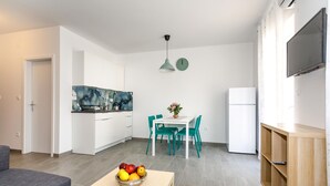 Una cocina moderna con armarios blancos, una mesa de comedor con sillas color turquesa, un frutero sobre una mesa de centro y un televisor montado en la pared.