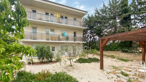 Un edificio de varios pisos con balcones, un área de jardín con grava y plantas, y una pérgola de madera.