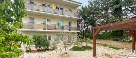 A multi-story building with balconies, a garden area with gravel and plants, and a wooden pergola.