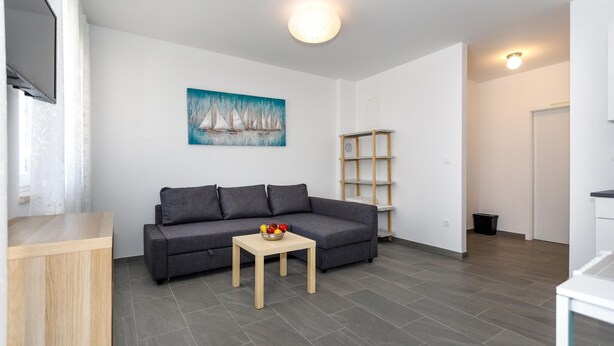Una sala de estar moderna con un sofá gris, una pequeña mesa de madera con fruta, un cuadro de veleros y una puerta blanca.