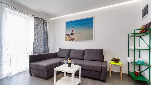 A living room with a grey sofa, a white coffee table, a painting of a lighthouse, a green shelf, and a window with sheer curtains. - Nice apartment in Krk with WiFi (Krk)
