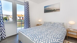 A bedroom with a bed, two bedside tables, a window with curtains, a painting on the wall, and a view of buildings outside. - Nice apartment in Krk with WiFi (Krk)