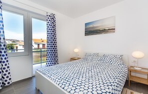 Ein Schlafzimmer mit einem Bett, zwei Nachttischen, einem Fenster mit Vorhängen, einem Bild an der Wand und Blick auf Gebäude.