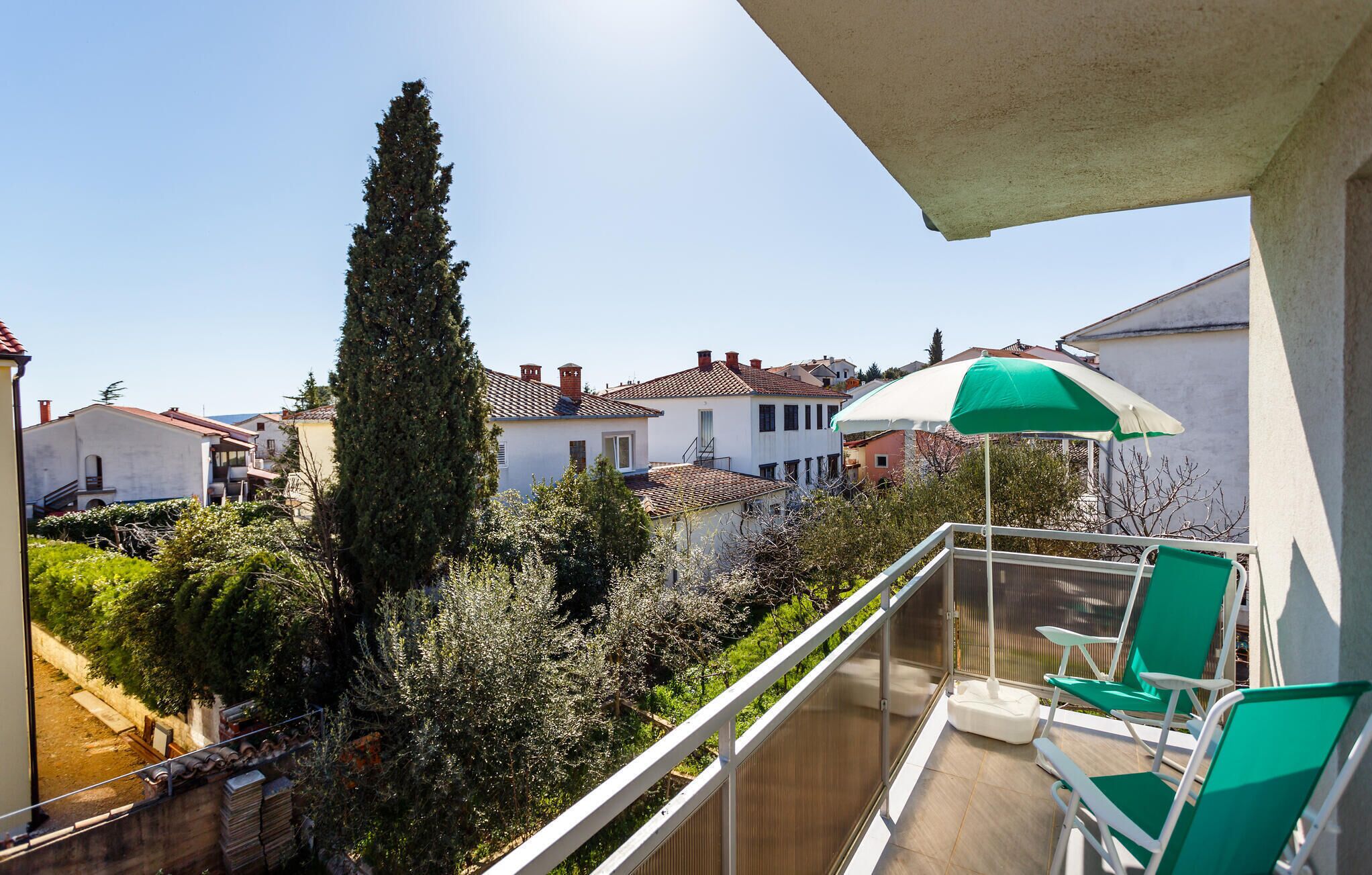 A balcony with a view of a residential area, a tall tree, and a green umbrella.