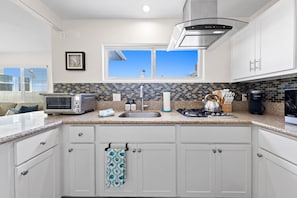 A modern kitchen with white cabinets, a stainless steel range hood, a window with a view, and a microwave on the counter. - The Tidal Retreat- Ocean Views (San Diego)