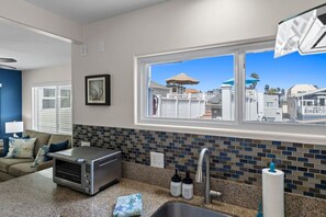 A kitchen with a toaster, a sink, and a window with a view of a white fence and umbrellas. - The Tidal Retreat- Ocean Views (San Diego)