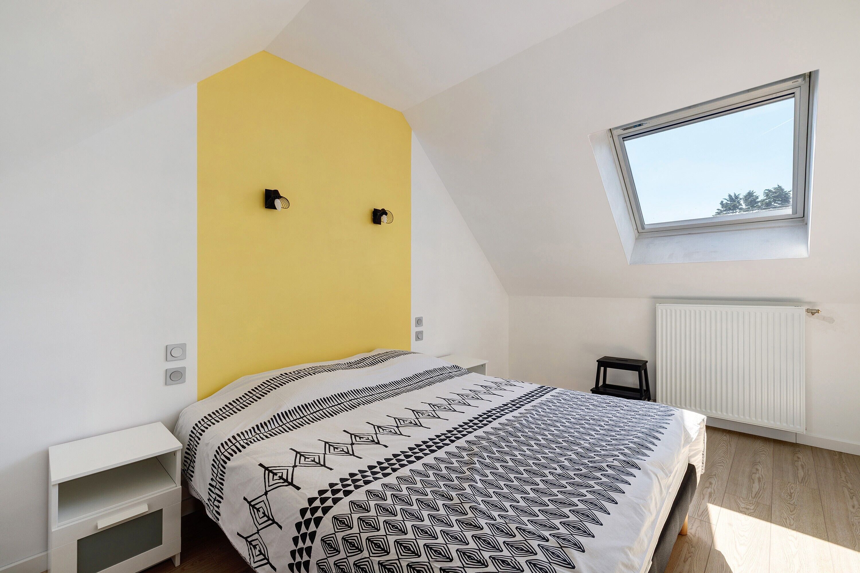 Een slaapkamer met een schuine kap, een raam, een bed met beddengoed met een patroon, een witte nachtkastje en een radiator.