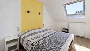 Une chambre avec un plafond en pente, une fenêtre, un lit avec une literie à motifs, une table de chevet blanche et un radiateur.