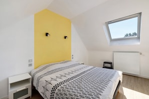 Ein Schlafzimmer mit schräg versiegelter Decke, einem Fenster, einem Bett mit gemusterten Textilien, einem weißen Nachttisch und einer Heizung.