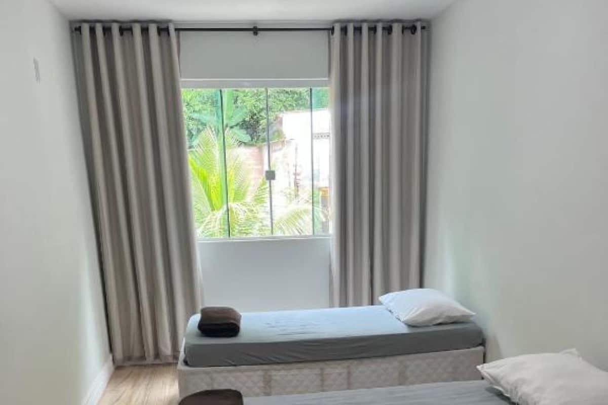 A room with two beds, a window with curtains, and a view of greenery outside.