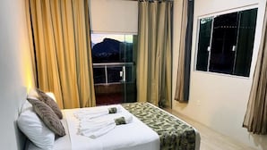 Una cama con una colcha blanca y estampada, una almohada y una toalla, con cortinas en las ventanas.