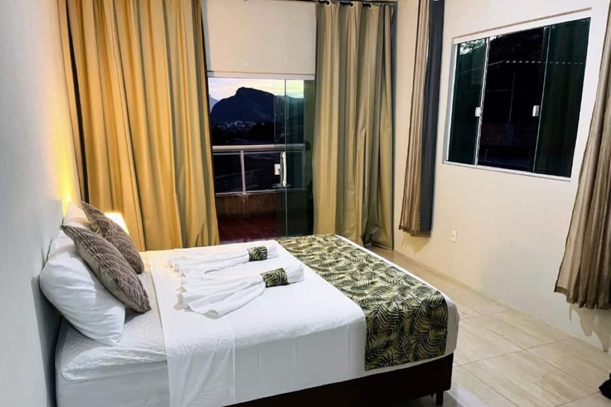 A bed with a white and patterned bedspread, a pillow, and a towel, with curtains on the windows.