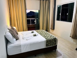 Una cama con una colcha blanca y estampada, una almohada y una toalla, con cortinas en las ventanas.