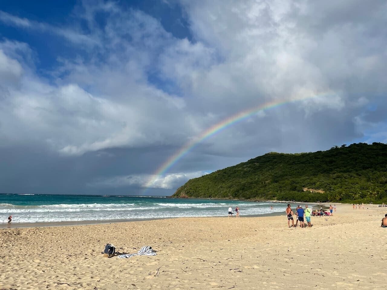 Una playa con arcoíris, orilla arenosa y una colina al fondo.