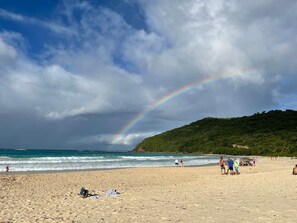 A beach with a rainbow, sandy shore, and a hill in the background.