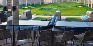 Ein Golfplatz mit Putting Green und zwei Golfwagen mit der Aufschrift 'TopGolf'. Im Vordergrund befinden sich Tische und Stühle.