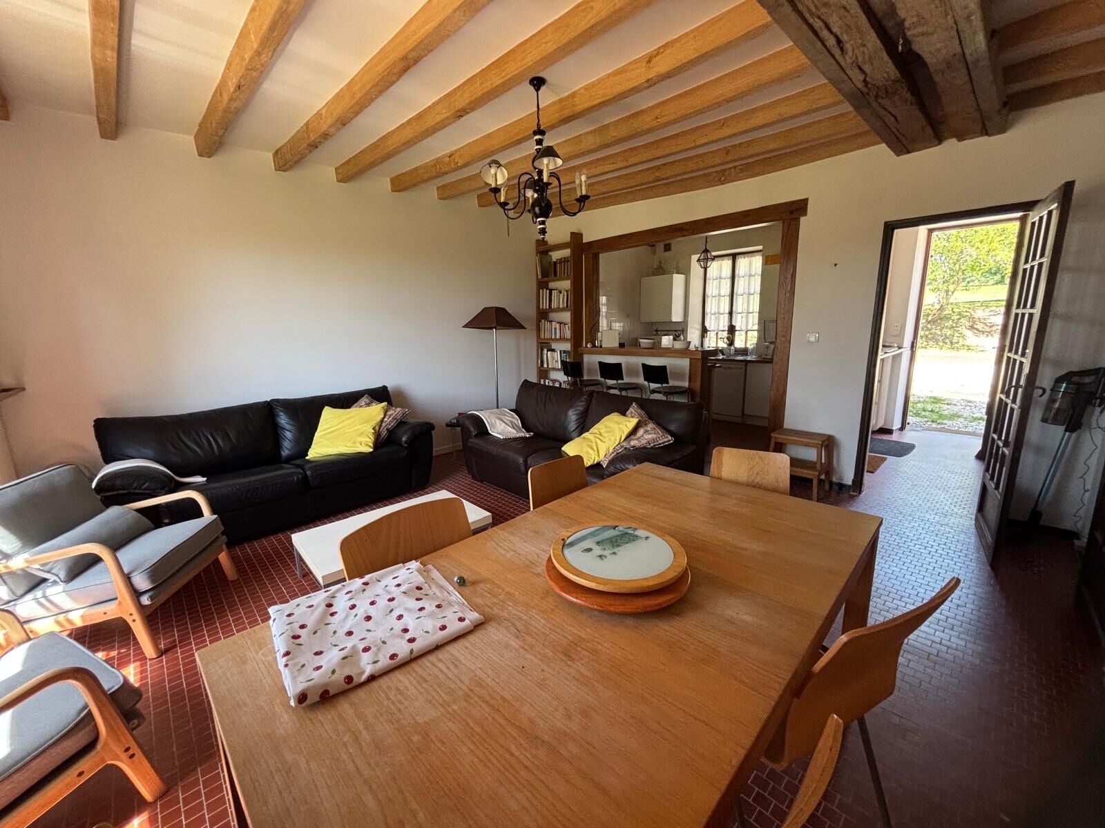 A living room with a wooden dining table, two black leather sofas, a bookshelf, and a kitchen area visible through an open door.