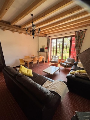 Ein Wohnzimmer mit einer Couch, einem Couchtisch und Sesseln. Es gibt einen Esstisch mit Stühlen, einen Fernseher und ein Fenster mit Vorhängen.