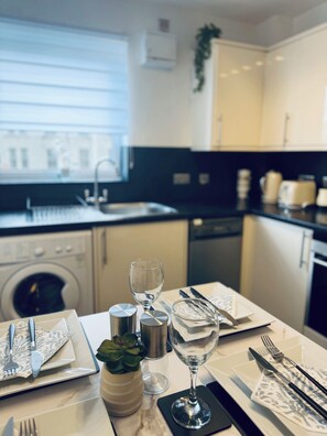 A modern kitchen with a dining area set for two, featuring a marble countertop, stainless steel appliances, and a view of the city. - Modern Central Aberdeen 2BR Apartment with Parking | Business & Leisure (Aberdeen)