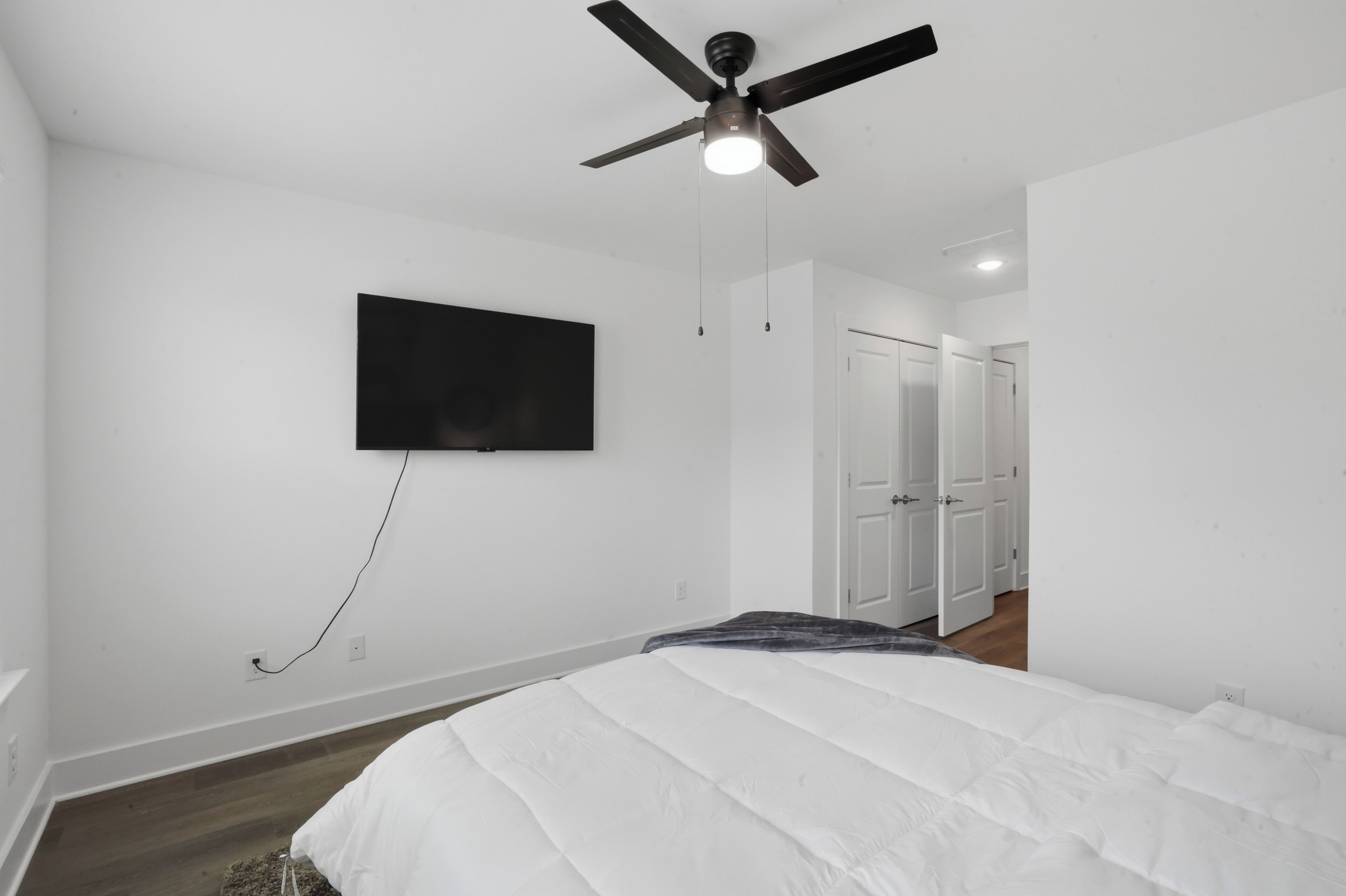 A bedroom with a bed, a ceiling fan, a TV mounted on the wall, and a door leading to another room.