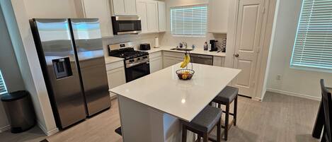 A modern kitchen with stainless steel appliances, a central island with stools, and a window with blinds.