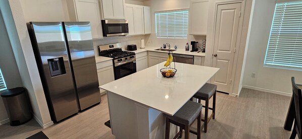 A modern kitchen with stainless steel appliances, a central island with stools, and a window with blinds.