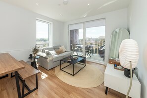 Ein modernes Wohnzimmer mit einer Couch, einem Couchtisch und einem Esstisch, großen Fenstern und einem Balkon mit Gartenmöbeln.
