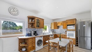 A modern kitchen with wooden cabinets, a washing machine, a dining table with chairs, and a large clock on the wall. - Gîte Cap with Terrace Near Beaches (Telgruc-sur-Mer)