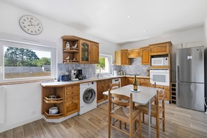 A modern kitchen with wooden cabinets, a washing machine, a dining table with chairs, and a large clock on the wall. - Gîte Cap with Terrace Near Beaches (Telgruc-sur-Mer)