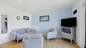 A modern living room with a grey sofa, a grey armchair, a TV on a stand, and two framed pictures on the wall. - Gîte Cap with Terrace Near Beaches (Telgruc-sur-Mer)