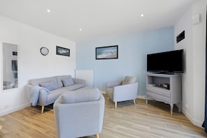 A modern living room with a grey sofa, a grey armchair, a TV on a stand, and two framed pictures on the wall. - Gîte Cap with Terrace Near Beaches (Telgruc-sur-Mer)