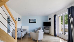 A living room with a staircase, a sofa, a TV, and a large window with curtains. - Gîte Cap with Terrace Near Beaches (Telgruc-sur-Mer)