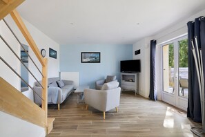 A living room with a staircase, a sofa, a TV, and a large window with curtains. - Gîte Cap with Terrace Near Beaches (Telgruc-sur-Mer)